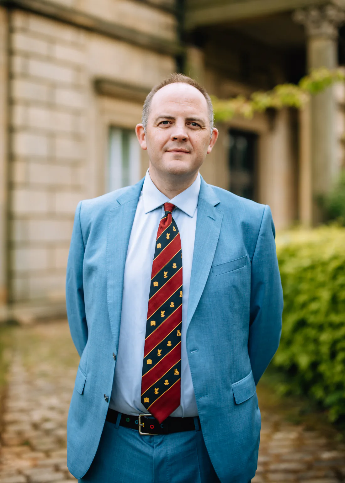 Mr Christopher West in a blue suit and red striped tie outside a stone building.