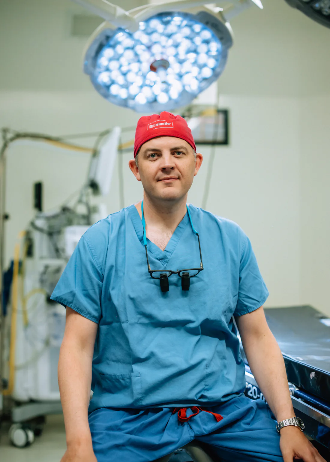 Mr Christopher West in blue scrubs, a red theatre cap and surgical loupes, sitting in an operating theatre.