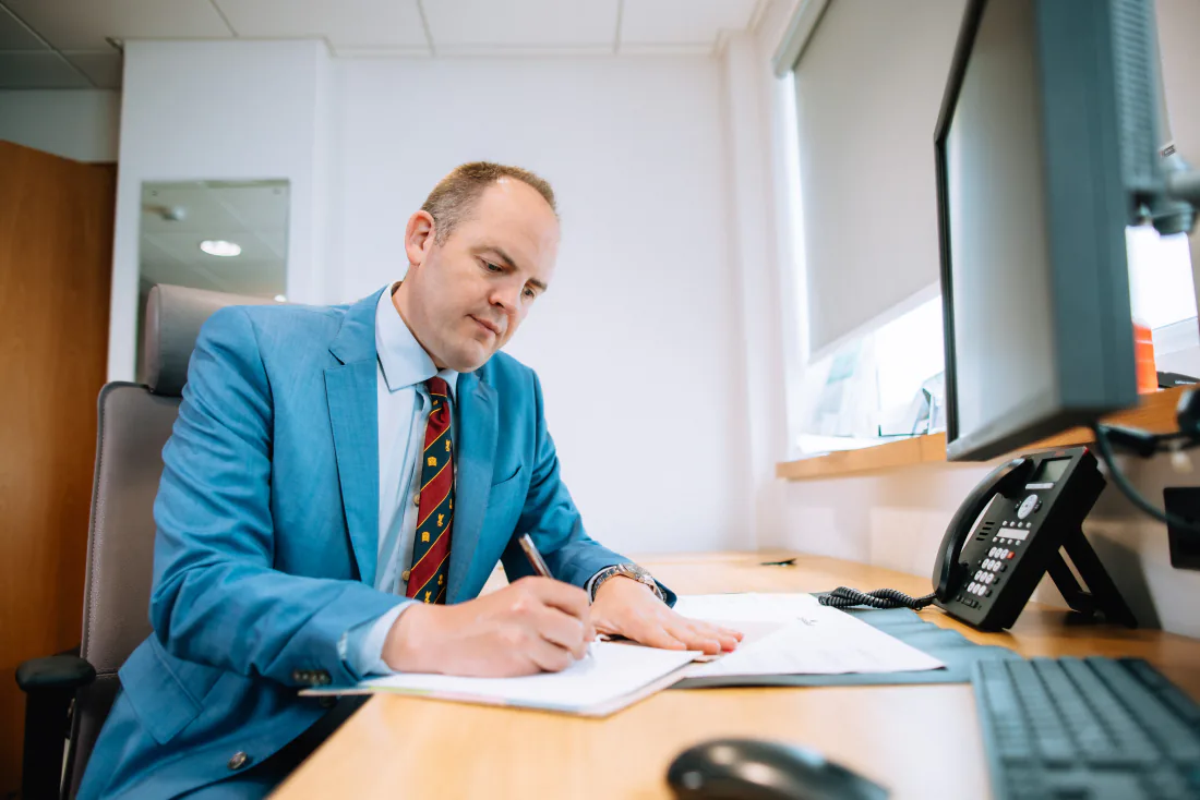 Mr Christopher West writing at his desk in a blue suit.