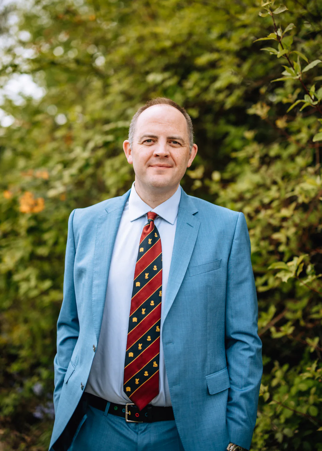 Mr Christopher West in a blue suit and striped tie outside a stone building.