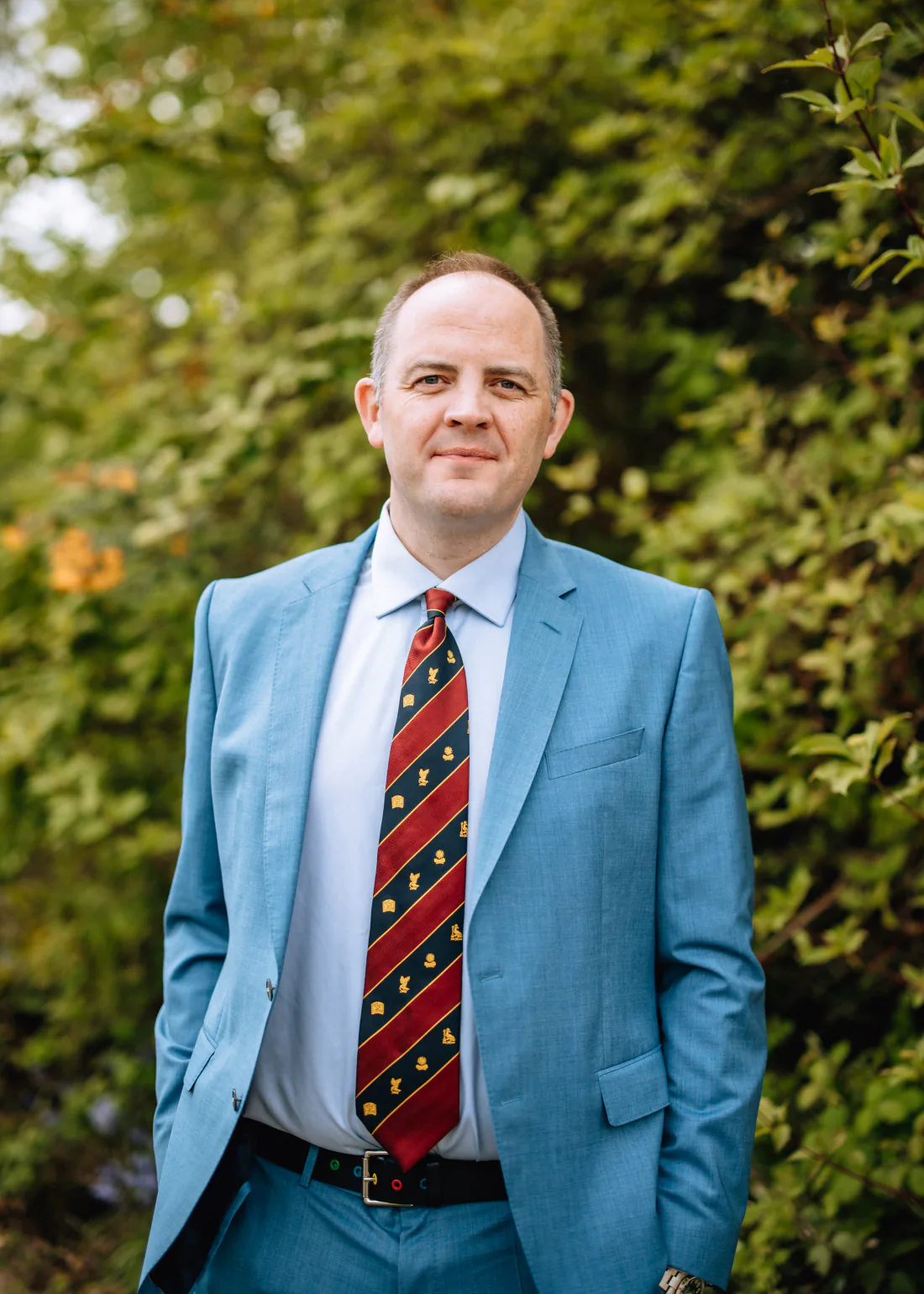 Mr Christopher West in a blue suit and striped tie outside a stone building.