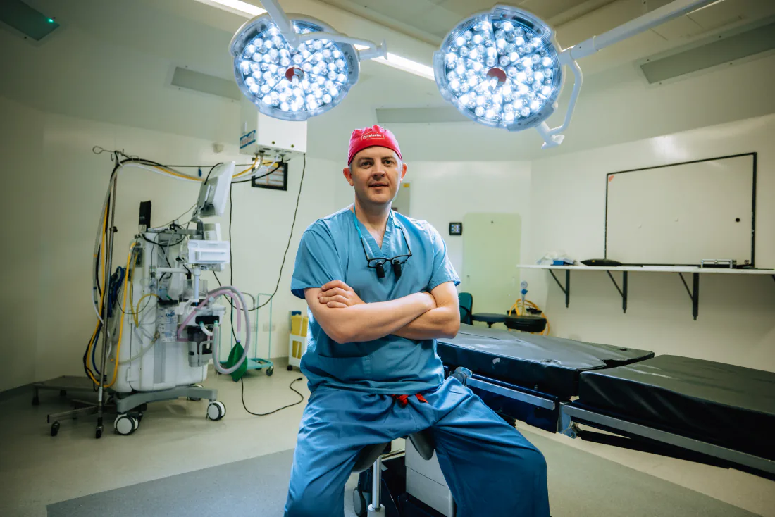 Mr Christopher West in blue scrubs and a red theatre cap in an operating theatre.