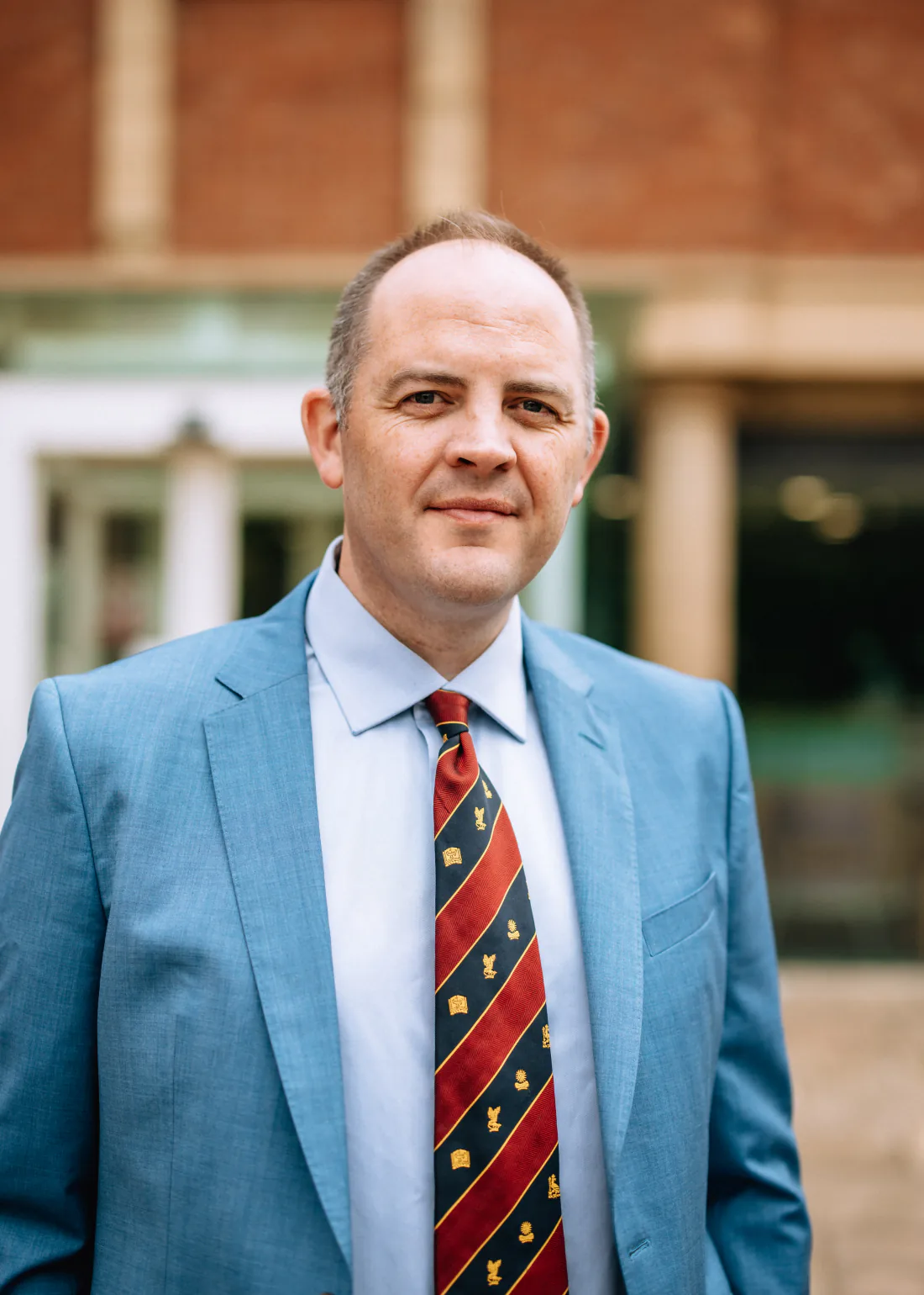 Mr Christopher West in a blue suit and striped tie outside a stone building.