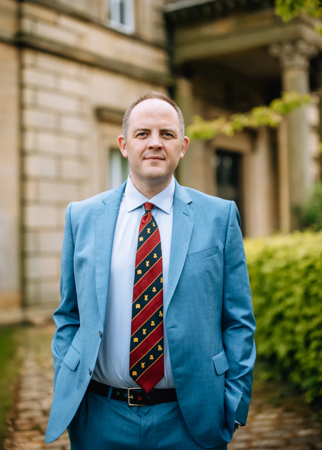 Mr Christopher West in a blue suit and striped tie outside a stone building.