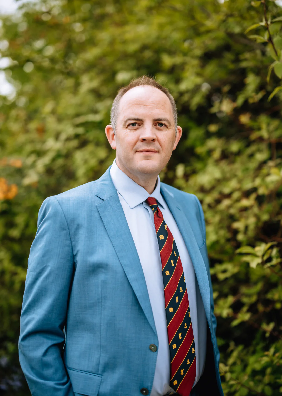 Mr Christopher West in a blue suit and striped tie, smiling, in front of green foliage.