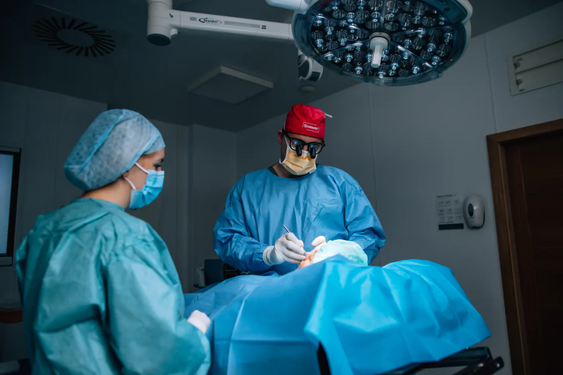 Mr Christopher West in a red cap and loupes at the operating table, a member of the theatre team beside him.