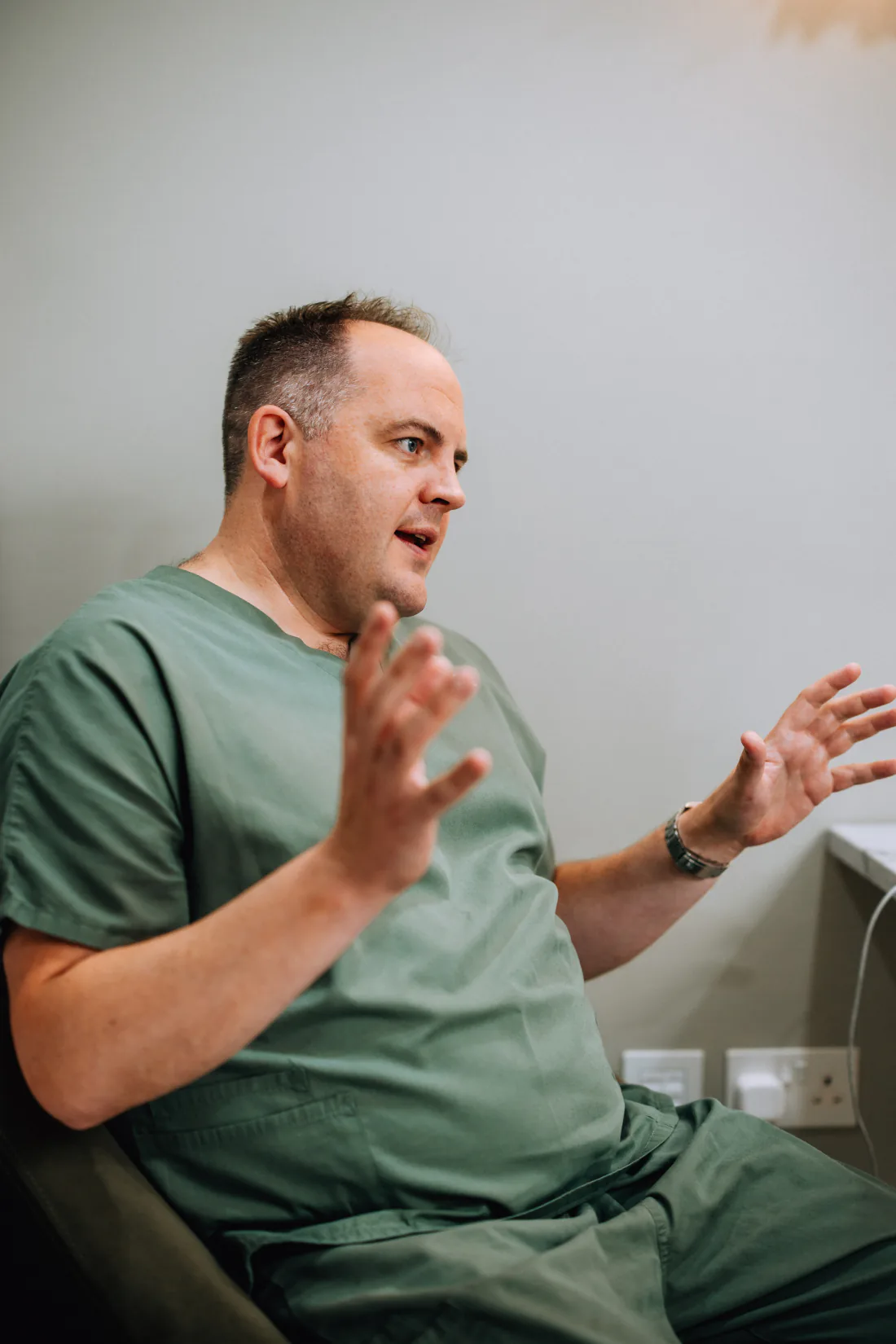 Mr Christopher West in green scrubs, seated and talking with both hands raised.