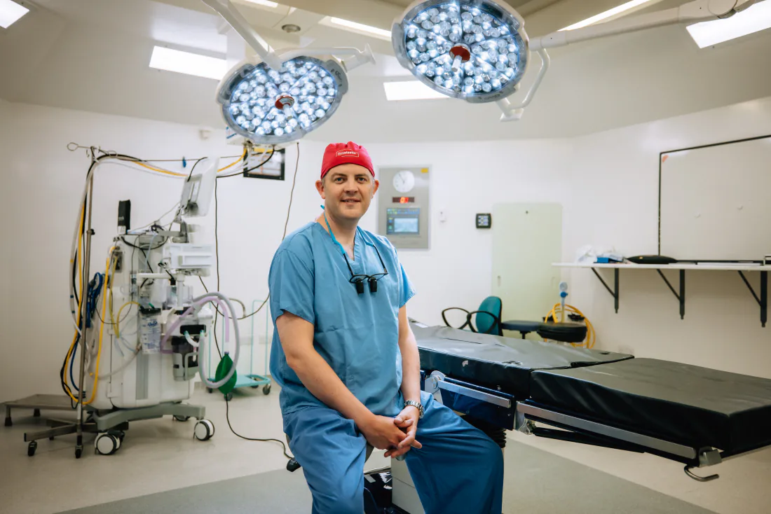 Mr Christopher West in blue scrubs and a red theatre cap in an operating theatre.