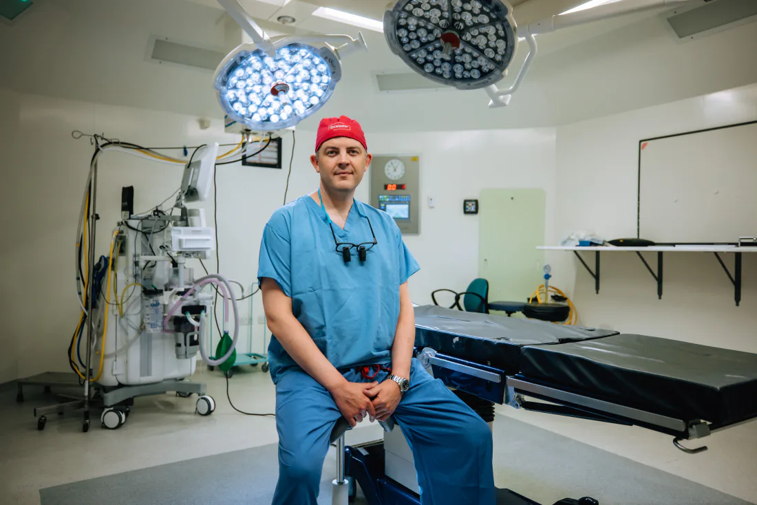 Mr Christopher West in blue scrubs and a red theatre cap, sitting by the operating table.