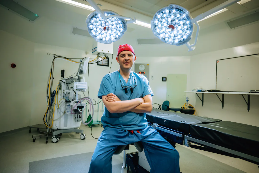 Mr Christopher West in blue scrubs and a red theatre cap, arms folded, in an operating theatre.
