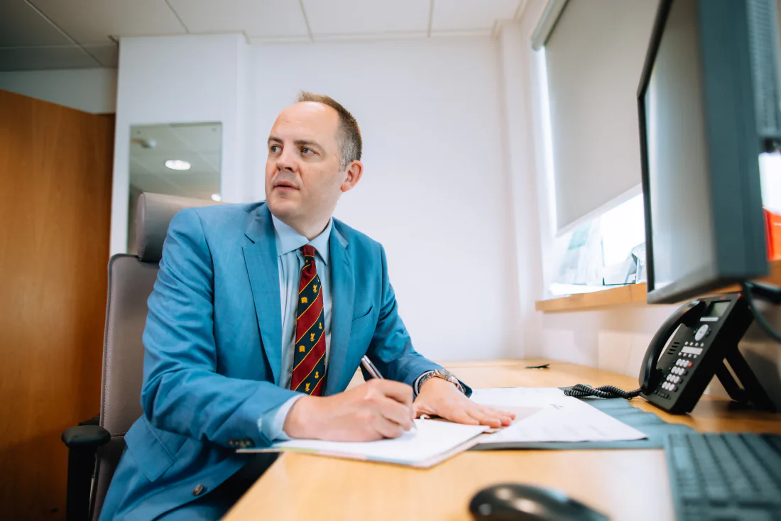 Mr Christopher West in a blue suit writing at his consulting room desk.