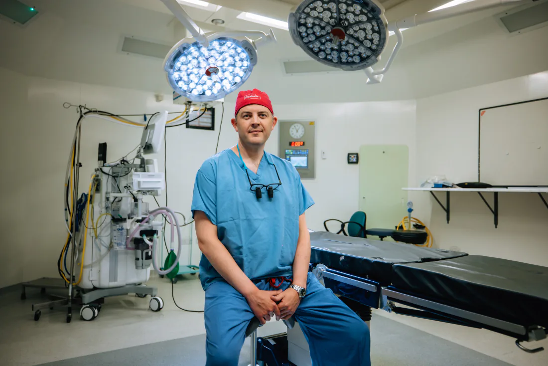 Mr Christopher West in blue scrubs and a red theatre cap in an operating theatre.