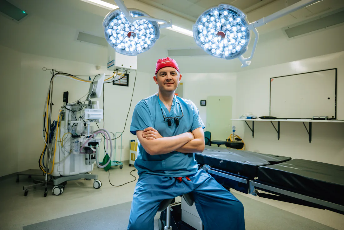 Mr Christopher West in blue scrubs and a red theatre cap in an operating theatre.