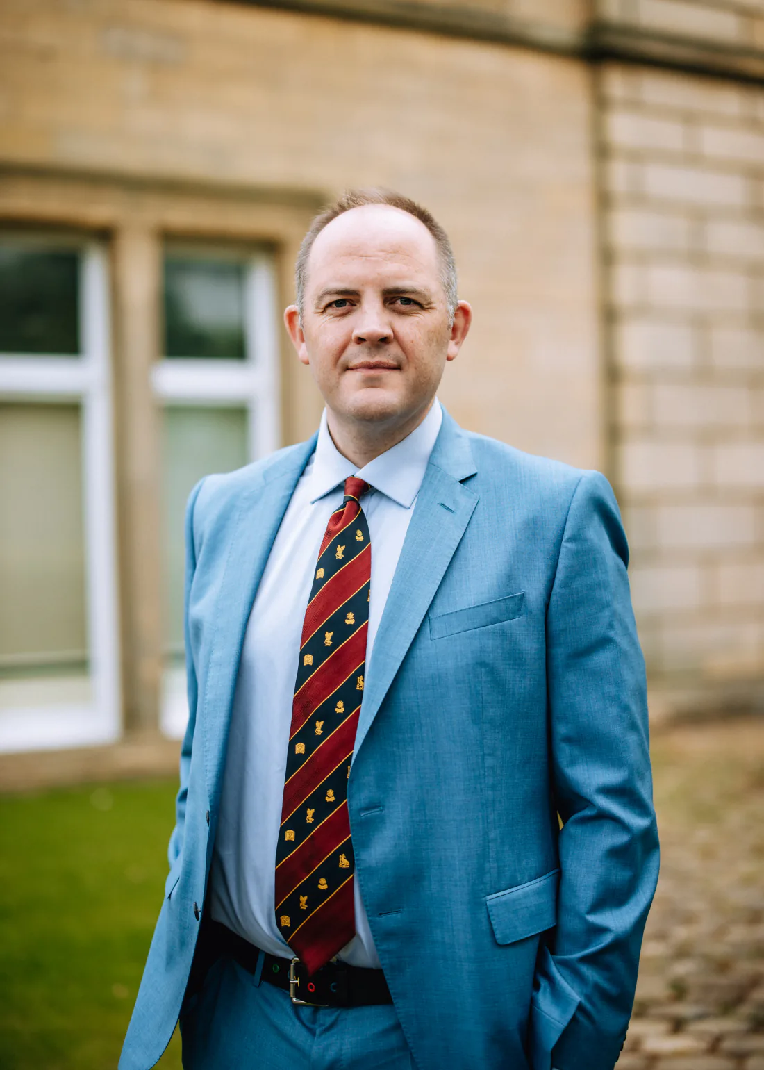 Mr Christopher West in a blue suit and striped tie outside a stone building.