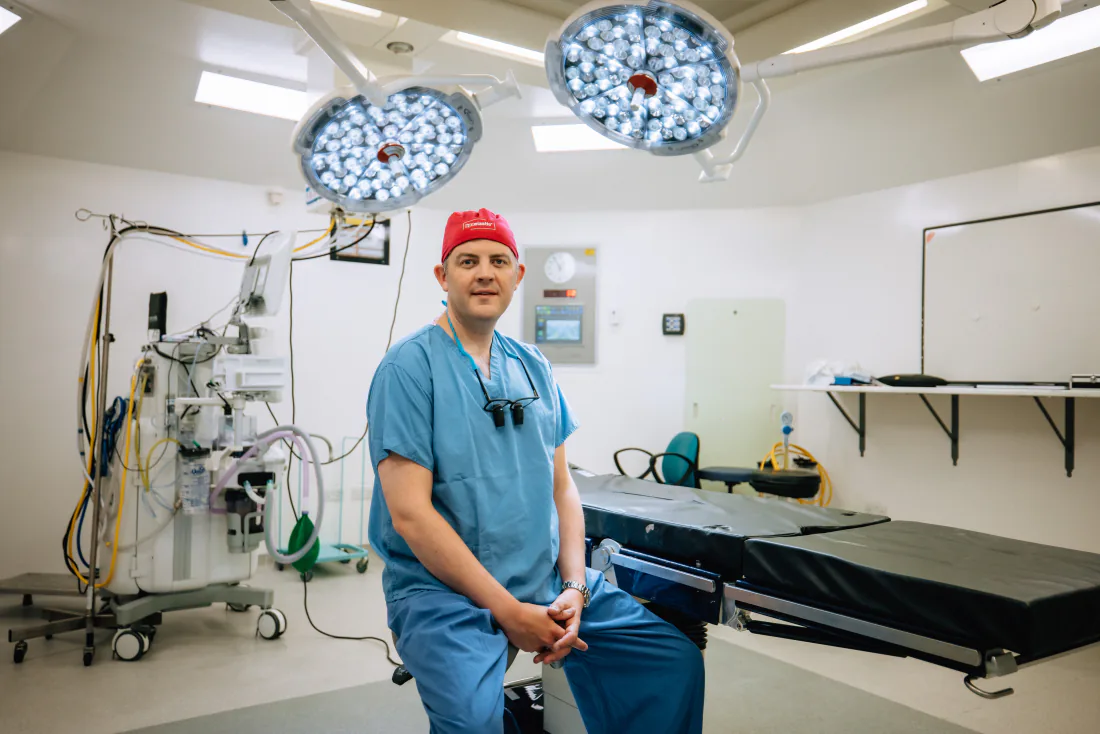 Mr Christopher West in blue scrubs and a red theatre cap in an operating theatre.