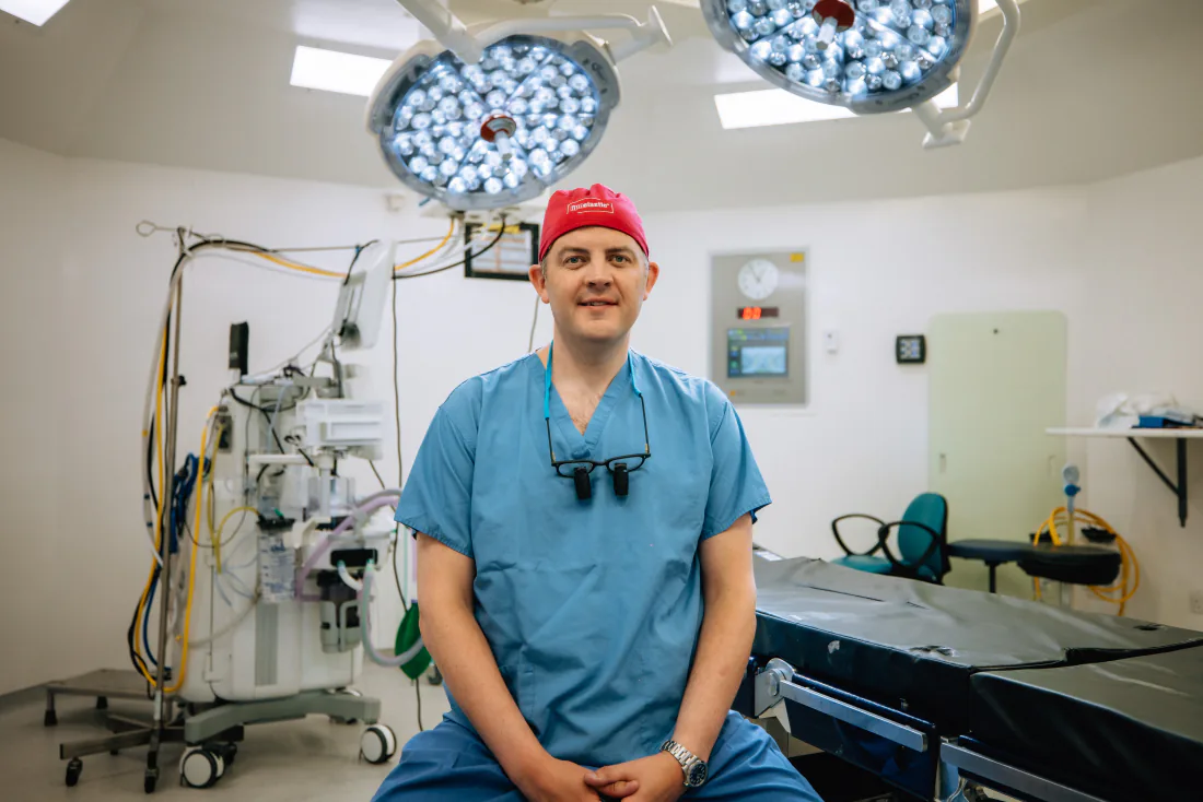Mr Christopher West in blue scrubs and a red theatre cap in an operating theatre.
