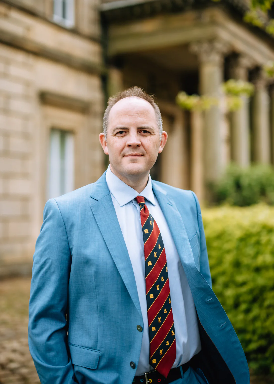 Mr Christopher West in a blue suit and striped tie outside a stone building.