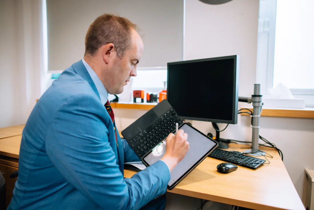 Mr Christopher West in a blue suit writing on a tablet at his consulting room desk.