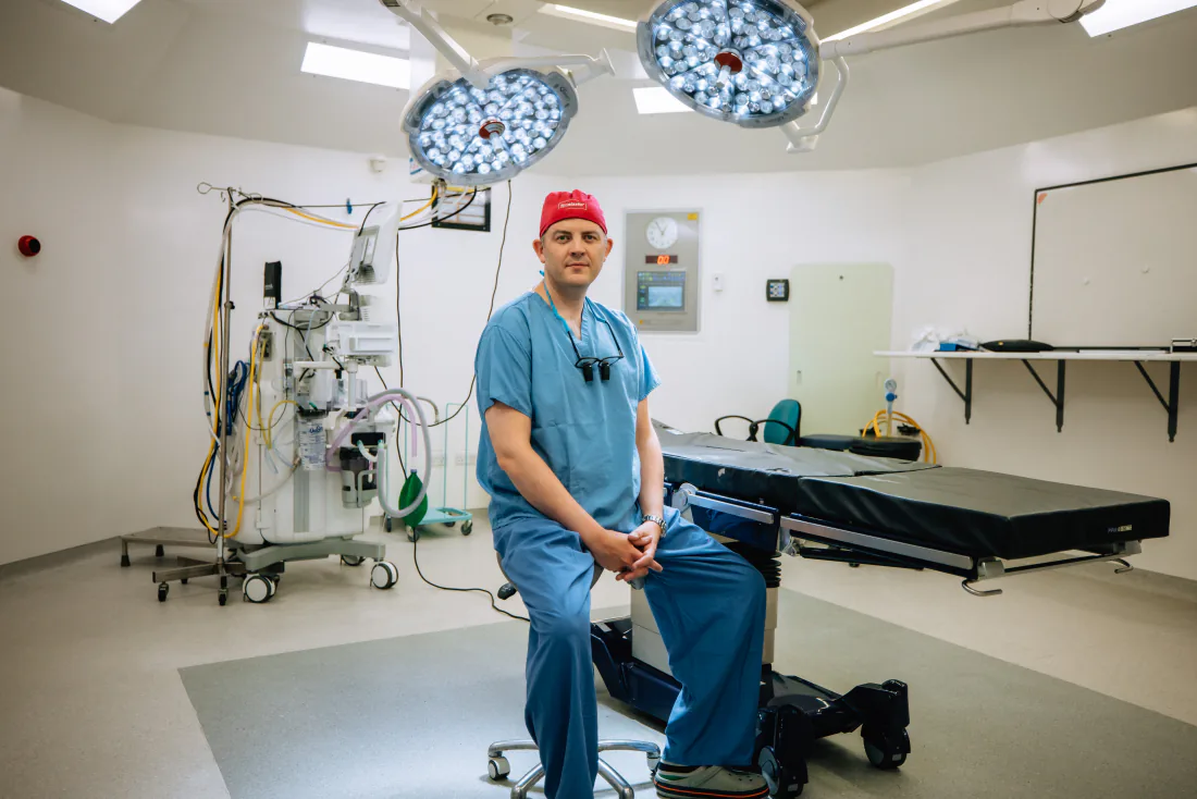 Mr Christopher West in blue scrubs and a red theatre cap in an operating theatre.
