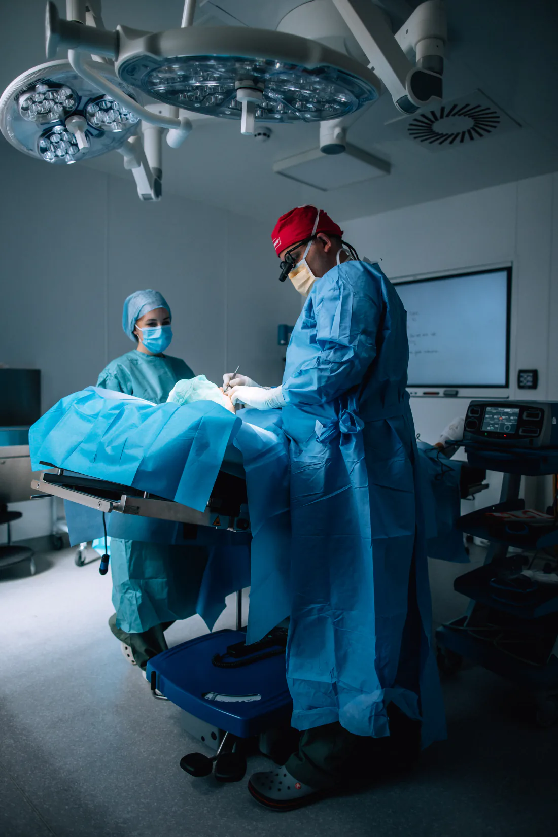 Mr Christopher West at the operating table in a surgical gown, red cap and loupes, a member of the team opposite.