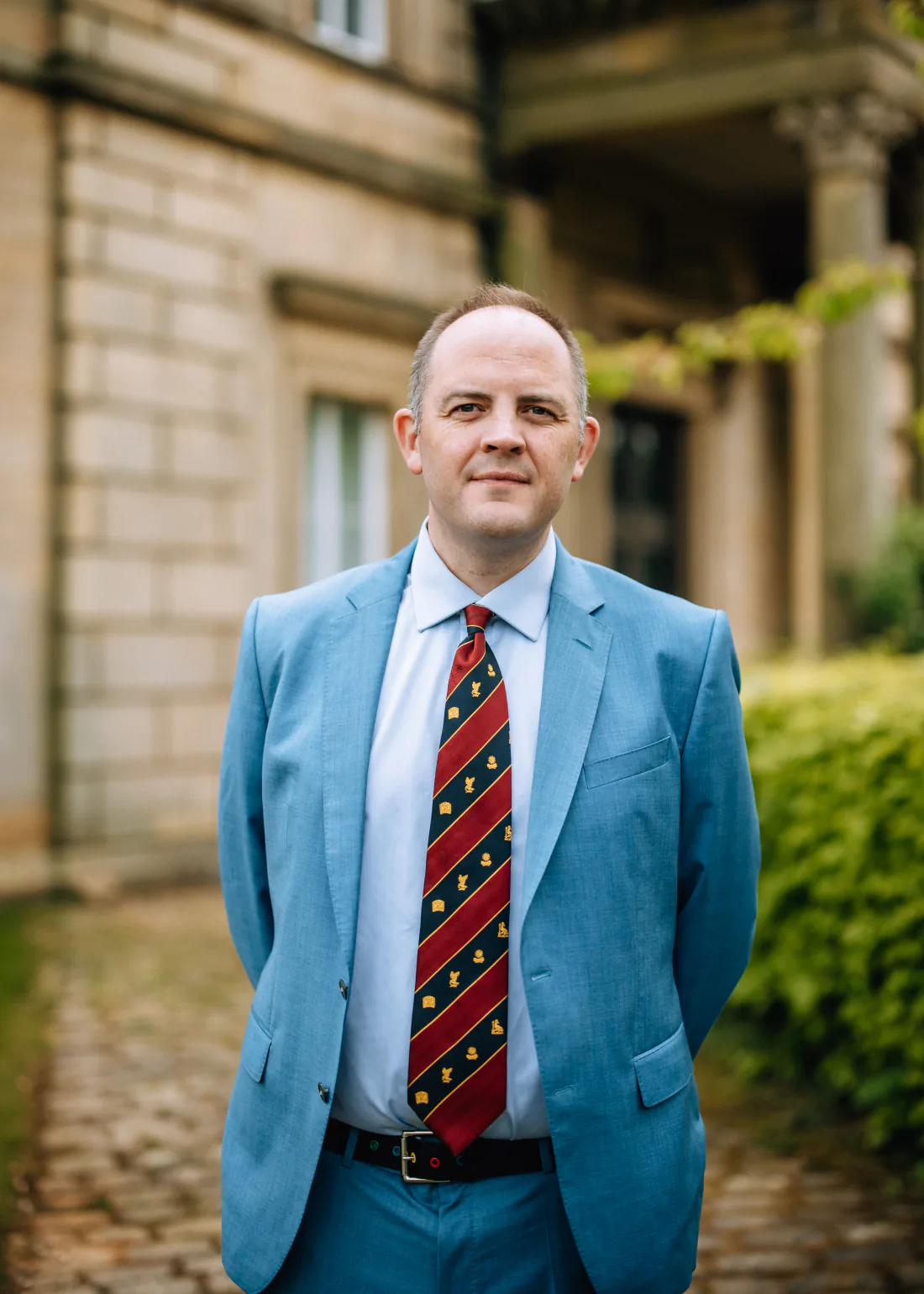 Mr Christopher West in a blue suit and striped tie outside a stone building.