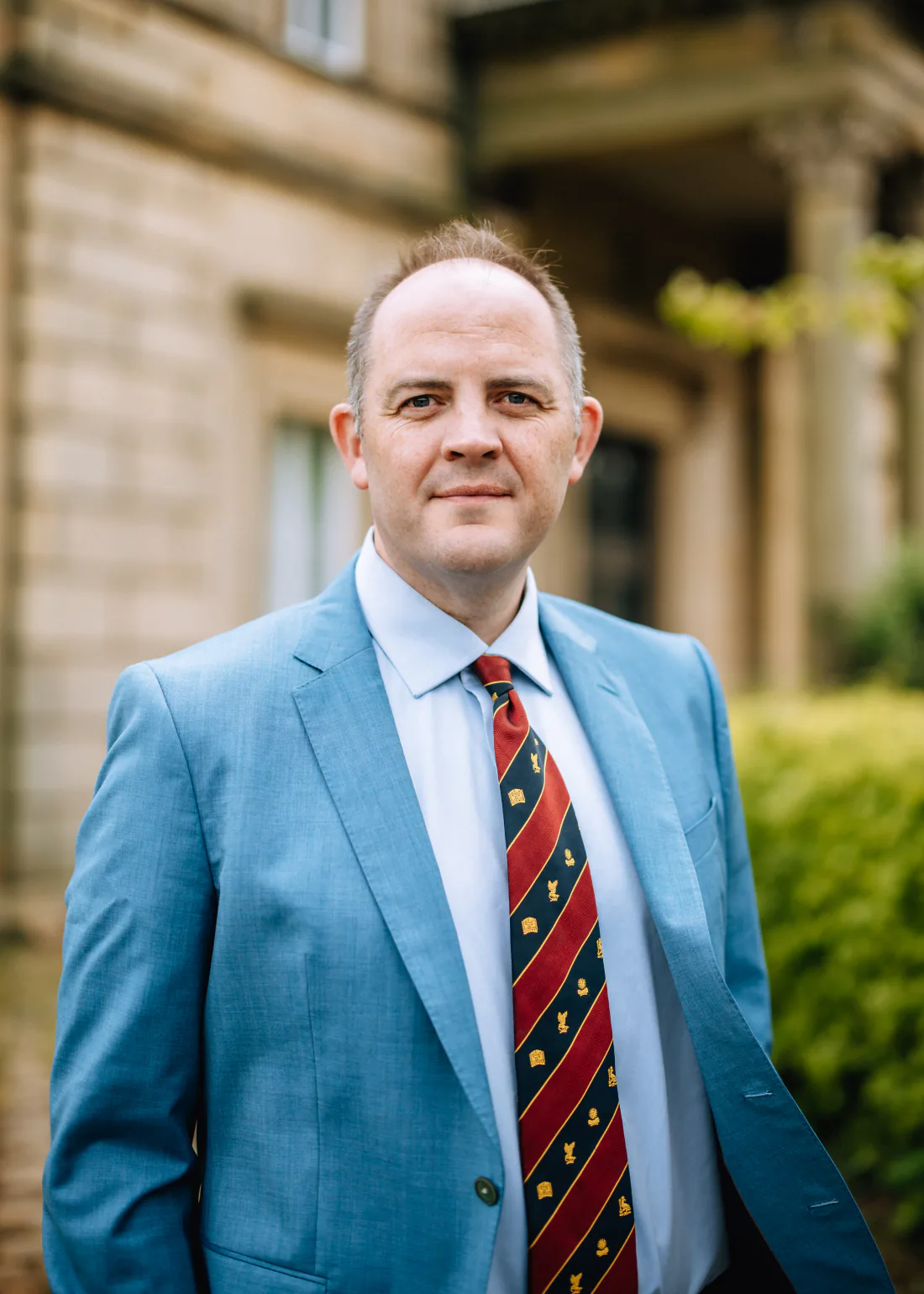 Mr Christopher West in a blue suit and striped tie outside a stone building.