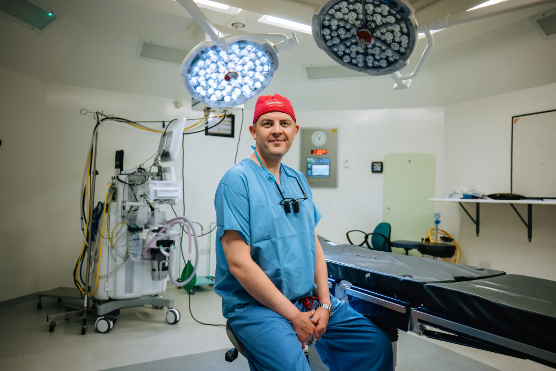 Mr Christopher West in blue scrubs and a red theatre cap in an operating theatre.