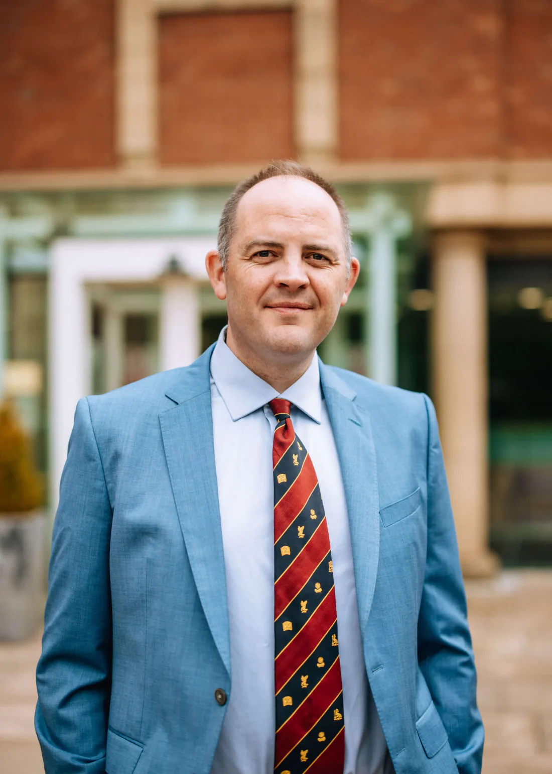 Mr Christopher West in a blue suit and striped tie, smiling, outside a red brick building.