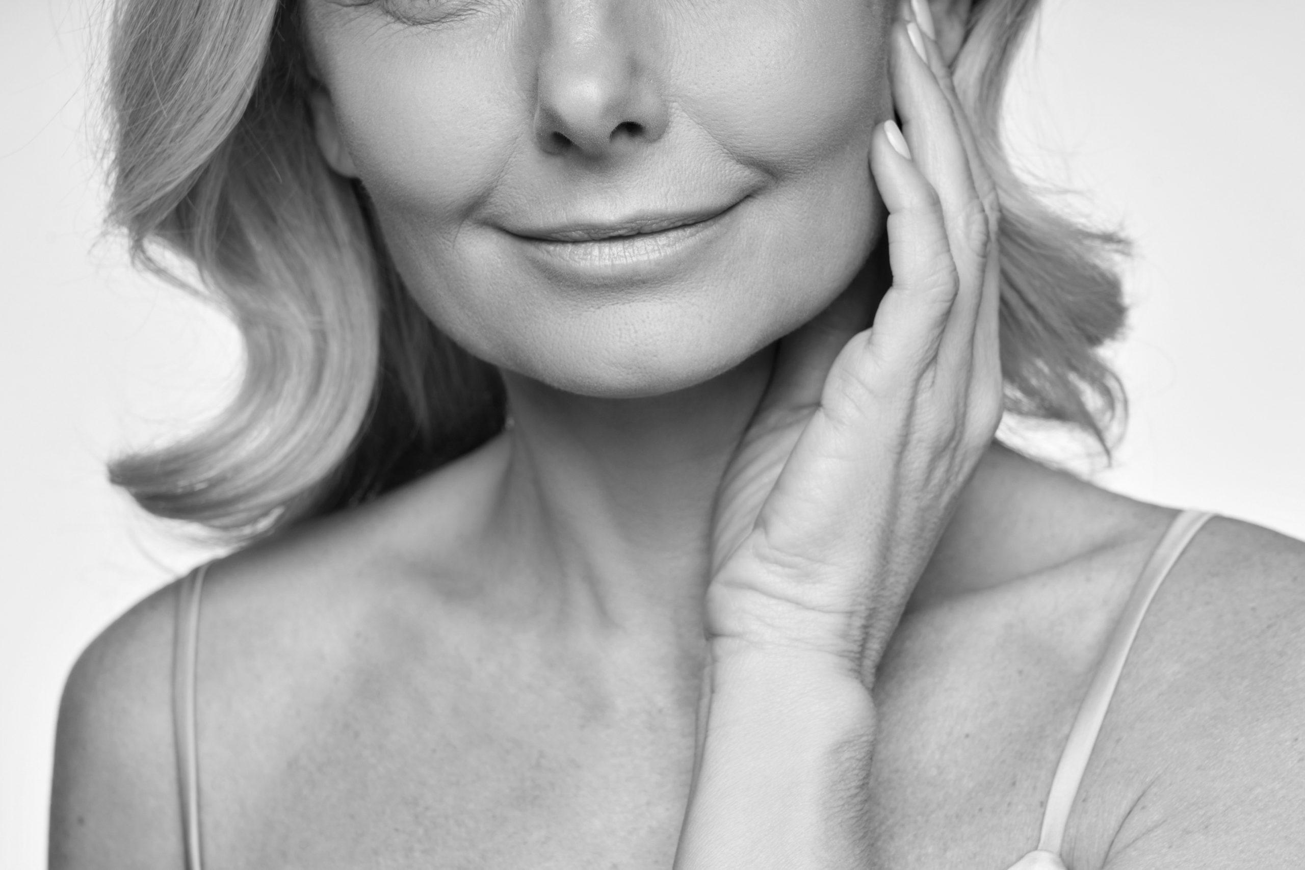 Black and white close-up of a smiling woman’s lower face and neck with her hand to her cheek